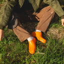 Person wearing a green jacket and brown pants with orange shoes sitting in grass