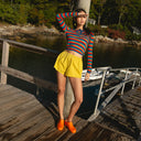 Woman in a striped sweater and yellow shorts and orange calzuro clogs standing on a wooden dock by a body of water.